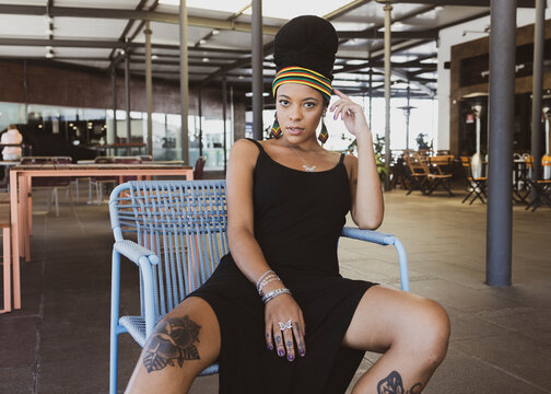 Beautiful tattooed African American woman wearing casual black dress staring seriously at the camera sitting on a chair inside a food court with foot resting on a small table. Copy space for text.