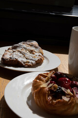 an almond croissant and a bun with strawberry slices lie on a white plate next to a white cup of coffee cappuccino and latte food fresh pastries bakery handmade kitchen cafe breakfast health