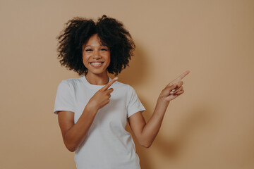 Positive cheerful dark skinned woman pointing at copy space isolated on beige background