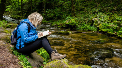 Naklejka na ściany i meble Young blond woman in a blue parka writing in her diary while sitting on a mossy rock by a fresh forest creek in mountains. Travel alone.