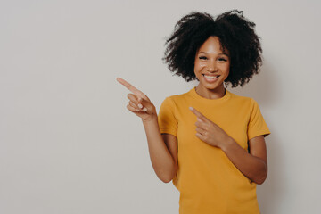 Cheerful dark skinned woman dressed in yellow tshirt pointing at left upper corner with forefingers