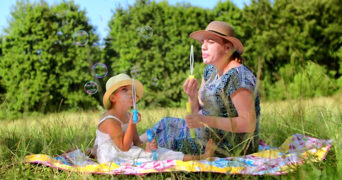 A Caucasian mother and her little five-year-old daughter play on the lawn in the forest and blow soap bubbles, have fun spending their leisure time and walking in the park in the summer.
