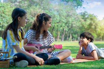 Two teen girl playing ukulele guitar while sitting on lawn green grass in city park, having fun with friends, boy lay on the lawn and listening to her music nearby