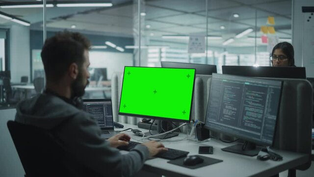 Multi-Ethnic Office: IT Programmer Working on Computer with Green Screen Chroma Key Display. Male Software Engineer Developing App, Program, Video Game. Terminal with Code Language. Over Shoulder Shot