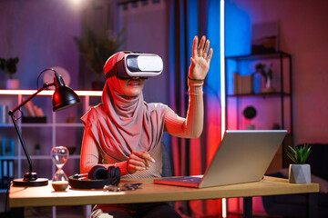 Modern smiling young Arab muslim woman in headscarf, using 3D augmented reality goggles at home in the evening, while working remotely on the project with imaginary screen, touching air