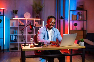 Handsome african man in casual clothes sitting at table and typing on wireless laptop. Young guy doing remote work on portable computer during evening time at home. Freelance concept.