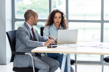Businessman and businesswoman sharing laptop while sitting at desk in office building room.