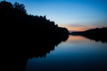 Sunset and its reflection on the water surface of the river. Bright colors and silhouettes of trees on the shore. Copy space