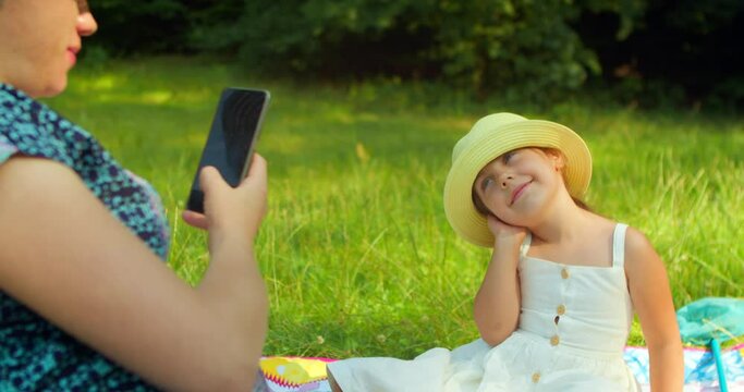 A pregnant mother takes pictures of her daughter on a smartphone against the background of trees sitting in a forest clearing. A little daughter poses for a photo in a beautiful hat.