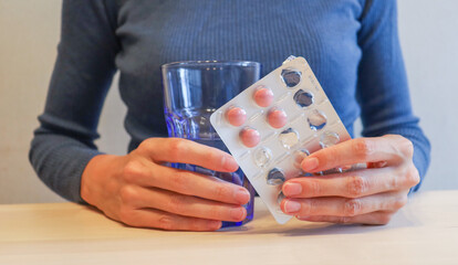 the girl is sitting at the table, holding pink pills in a package and a glass of water in her hands. Lack of vitamins in autumn and winter, uncontrolled medication