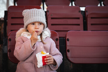 Cute little caucasian girl eating French fries at the stadium while watching a football game. Beautiful little girl in a stylish outfit © alexanderon
