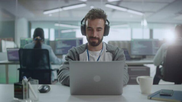 Office: Portrait of Smiling IT Programmer Wearing Headphones Working on Desktop Computer. Male Website Developer, Software Engineer Developing App, Video Game. Listening to Podcast, Music. Front View