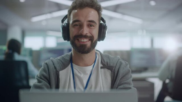 Office: Portrait of Happy IT Programmer Wearing Headphones Working on Desktop Computer, Looking at Camera and Smiling. Male Software Engineer Developing App, Video Game. Listening to Podcast, Music