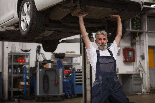 Bearded senior car service mechanic supporting automobile on car lift, making funny face