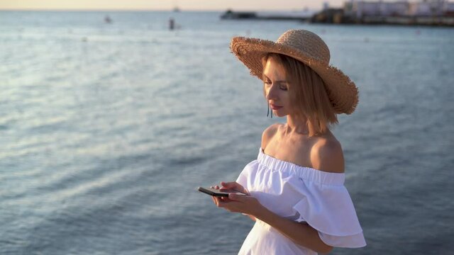 A young Caucasian woman walks near the sea or ocean in the early morning at dawn, holding a smartphone in her hands, leafing through the news feed. copy space. 4k