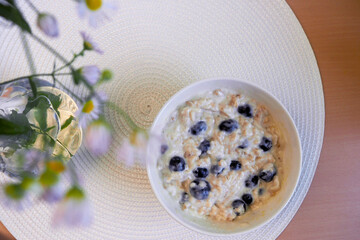 in a round white plate with oatmeal porridge with coconut milk and blueberries, which lies on a wooden table with a white rug next to chamomile flowers . top view