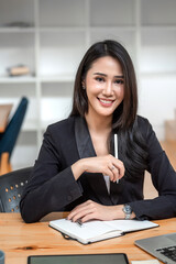 Charming young Asian businesswoman holding pen sitting at her desk with laptop notebook. Looking at camera.