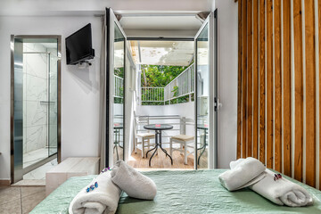 Exit from bedroom to the balcony with table and two chairs throught open french window. Towels on the bed, TV on the wall. White colors with the wooden elements.