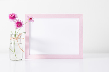 Pink frame mockup on white wall with pink flowers in glass vase decoration. Front view