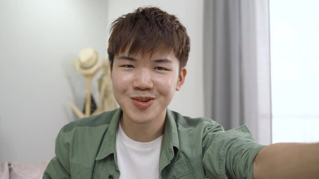 headshot happy asian young man facing phone camera is talking with a bashful smile during a virtual chat online in the living room at home.
