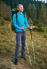 Full length of male hiker with backpack holding trekking poles and smiling while standing on grassy hill. Young happy man traveler enjoying beauty of nature while hiking alone in mountains.