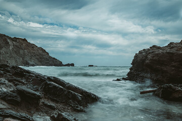 Fototapeta na wymiar rocky beach landscape during storm with cloudy sky. Dramataic long exposure of sea waves breaking on rocks.