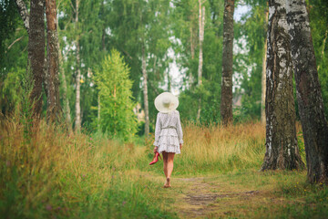 a woman in a short white dress and a hat walks barefoot  along a path among tall grass and trees with shoes in her hands