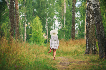 a woman in a short white dress and a hat walks barefoot  along a path among tall grass and trees...