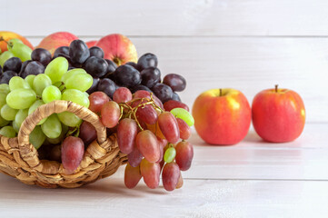 Different varieties of grapes and apples in a wicker basket on a light wooden background. Copy space.