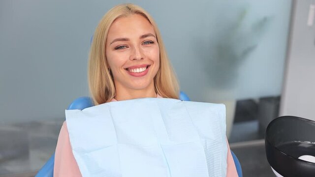 Satisfied smiling blonde young woman patient show thumb up like gesture sitting in chair in dental clinic light office medical center with modern tools equipment. Healthy lifestyle treatment concept