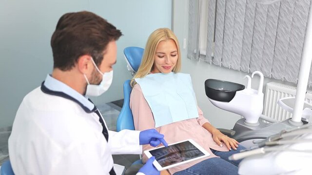 Male professional dentist doctor man shows an X-ray photo of teeth jaws on tablet pc computer to young woman patient work in dental clinic in light office medical center with modern tools equipment