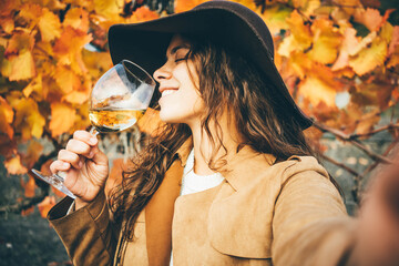 Smiling young woman in stylish hat and coat drinks delicious white wine from wineglass and making...
