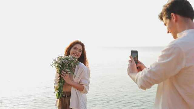 Young couple two friends family man woman 20s in white clothes hold flowers bouquet take photo on mobile cell phone rest at sunrise over sea beach ocean outdoor seaside in summer day sunset evening