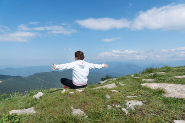 Naklejka na ściany i meble Young female with arms outstretched is sitting on top of mountain peak with sense of freedom. Copy space, healthy life and relaxation concept