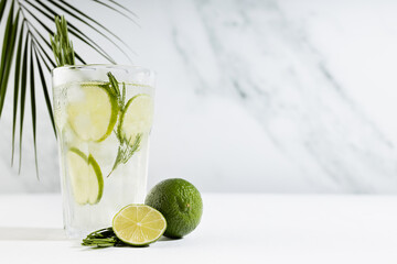 Refreshing  mojito cocktail with ice cubes, lime slices, green rosemary twig, ingredients, tropical palm leaf in elegant modern white interior on soft light wood table.