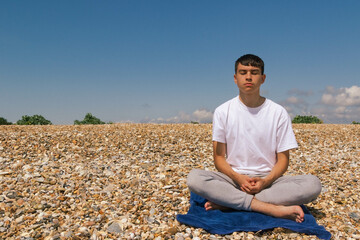 Fototapeta na wymiar A Caucasian teenage boy meditating on a stony beach with his hands resting on his lap