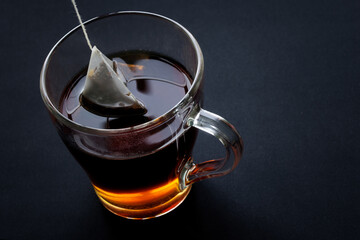 Black tea in a pyramid tea bag is brewed in hot water in a transparent cup. Black background. Diagonal top view. Free space for an inscription