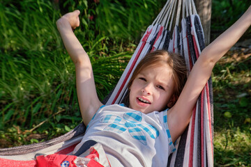 A happy little girl lies in a hammock in the garden in the summer. A child in a hammock during the holidays smiles in the park against the background of green grass. High quality photo