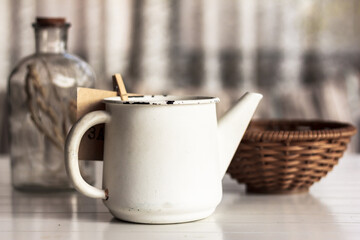 Kitchen décor, café vintage décor. White rustic watering pot, a decorative glass bottle and a basket, on white wooden table © Anna