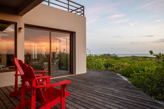 wooden deck terrace with two red chairs overlooking the beach, vegetation and ocean with the sunrise reflecting in the windows of an oceanfront luxury vacation home