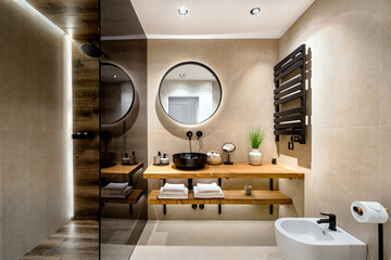 Modern tiled bathroom in beige and brown warm colors.  It has shower cabin with glass partition, wooden stand for black sink and round mirror on the wall. 