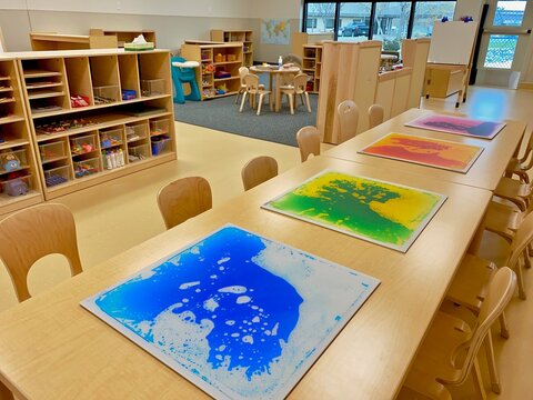 Early childhood education preschool classroom. Tables with art, chairs, and wooden shelves with supplies in clear bins. No people. Oil and water mix in squares. 