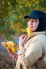 A Muslim woman in a hijab in a beige trench coat stands with autumn leaves. Close up. Black cap.