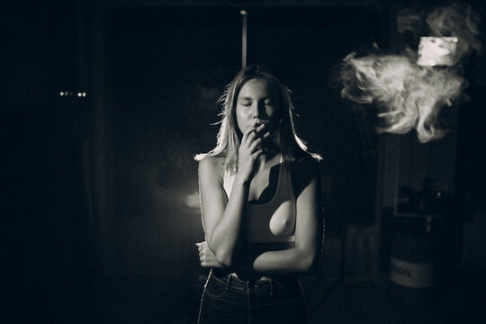 Young woman smokes cigarette in the dark with light spot ray. Black and white image with film grain