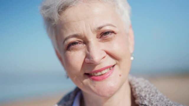 Closeup portrait of senior woman with gray short hair and makeup looking at camera and smiling on beach. Mature female posing on sea coast. Travel, aging and happy retirement concepts