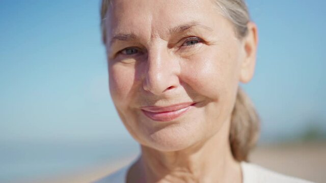 Closeup portrait face of beautiful senior woman with gray hair looking at camera and smiling outdoors against blue sky. Beauty, aging and happy retirement concepts