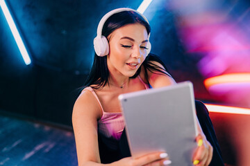 Modern devices. Beautiful young woman working on tablet with wireless headphones in neon light