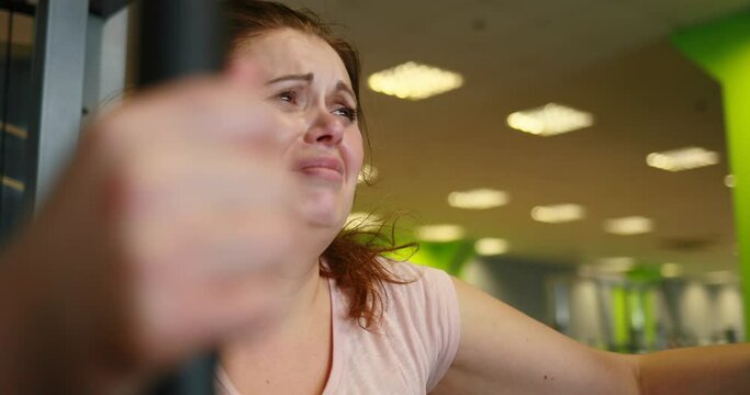 The depressed almost crying obese woman is hardly gaining weight on the pec deck training machine in green gym.