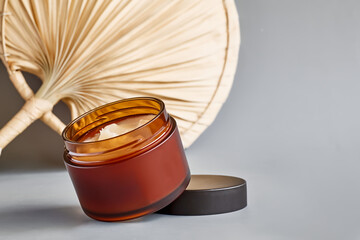 Open brown cream jar near dried palm leaves on pastel gray background. Product presentation....