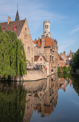 Brugge, Flanders, Belgium - August 4, 2021: Sunlit Belfry towers over brown brick back facades, mirrored in quiet Dijver canal, with green foliage in corners under blue sky. 